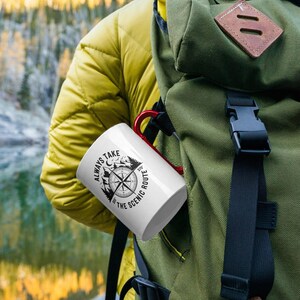 May include: White ceramic mug with a red handle featuring a black and white compass design and the text "ALWAYS TAKE THE SCENIC ROUTE". The mug is held in a green backpack pocket, suggesting outdoor or travel use.
