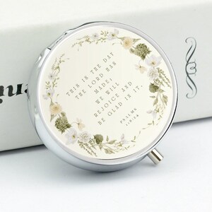 May include: A round silver compact mirror featuring a floral wreath design in soft green and white tones. The center displays the text: This is the day the Lord has made; we will rejoice and be glad in it. Psalms 118:24, printed in black.
