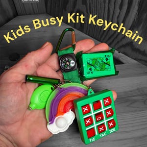 May include: A green keychain featuring a compass, a deck of cards, a rainbow, a whistle, and a Tic-Tac-Toe game. The keychain is labelled "Kids Busy Kit Keychain". The items are in various colours, including green, red, purple, and white.