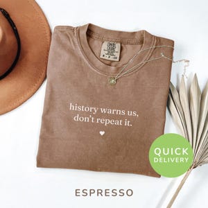 May include: Espresso-colored t-shirt with the text "history warns us, don't repeat it." and a small heart. A gold pendant necklace and a brown hat are next to the shirt. The shirt has a crew neck and short sleeves.