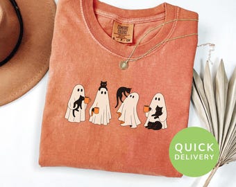 May include: Orange Comfort Colors t-shirt with a crew neck. The shirt features a graphic of four cartoon ghosts with black cats, holding coffee mugs. The word "YAM" is printed at the bottom. A gold necklace is draped on the shirt.