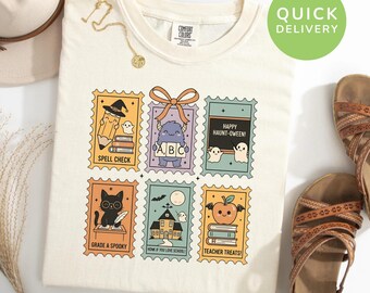 May include: Ivory-colored t-shirt featuring six colorful stamp-like designs with Halloween and school-themed illustrations. Designs include a witch's hat, ghosts, a monster, a black cat, a haunted house, and an apple. Text includes "Spell Check" and "Teacher Treats!"