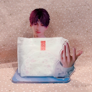 May include: A clear acrylic stand featuring a stylized illustration of a person with purple hair and a hand gesture. The stand holds a white package with pink floral accents and red text.