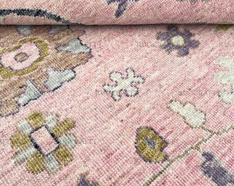 May include: Close-up of a rolled-up pink rug with a floral pattern. The rug features a light pink background with various floral and leaf designs in shades of purple, green, and white. The rug has a textured appearance.