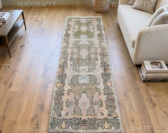 May include: A long rectangular runner rug featuring a repeating botanical pattern in muted sage green, soft pink, and cream tones. The design includes stylized leaf motifs and floral elements arranged symmetrically along the length of the rug.
