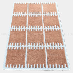 May include: A rectangular rug with a repeating pattern of peach-colored blocks separated by white lines. The blocks have a unique, textured design. The rug's edges are bordered with white. This rug is suitable for home decor.
