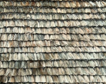 Puede incluir: Una alfombra texturizada con filas horizontales de material fibroso en capas, en tonos bronceado, beige y gris apagado. La superficie consiste en densos mechones superpuestos, creando una apariencia peluda y escalonada en toda la pieza.