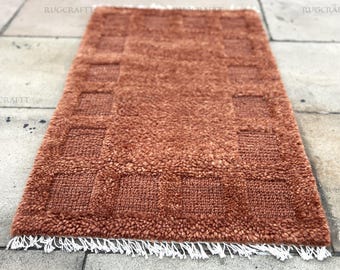 May include: A rectangular, rust-colored rug with a textured, plush surface and a geometric pattern of squares. The rug has a fringe of white tassels along one edge. The rug is displayed on a stone tile surface.
