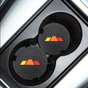 May include: Two black silicone cup holders with a mountain range design in yellow, orange, and red. The cup holders are in a car's cup holder.