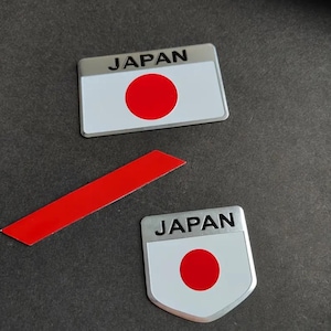 May include: Two Japanese flag-themed stickers. One rectangular sticker and one shield-shaped sticker, both with a white background and a red circle. The word "JAPAN" is printed in black on a silver bar at the top of each sticker.