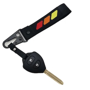 May include: Black car key with a metal key blade attached to a black fob. The key is connected to a black strap with a metal clasp. The strap has a colorful design of red, orange, and yellow diamonds.
