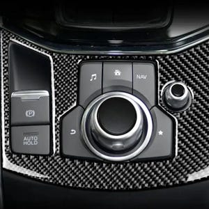 May include: Close-up of a car's center console with a carbon fiber pattern. Features buttons for music, home, and navigation. Includes a dial and buttons for parking and auto hold.