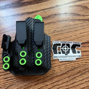 May include: A black and green gun holster with a carbon fiber pattern and green screws. The holster has a clip on the back and a logo that says "CLOSE QUARTERS CARRY" and "WWW.CLOSEQUARTERS CARRY.COM".