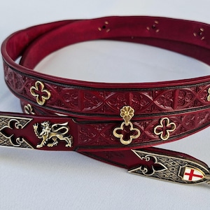 Peut inclure: Une ceinture en cuir bordeaux avec des motifs floraux et géométriques en relief. La ceinture présente une boucle argentée et des détails décoratifs, dont un emblème de lion doré et des détails de fleur de lys. La ceinture mesure environ 3,8 cm de large.