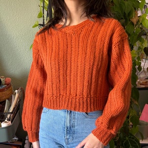 May include: A cropped orange knitted sweater with long sleeves and a ribbed texture.