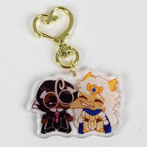 May include: A gold keychain with a heart-shaped loop and a clear acrylic charm featuring two cartoon characters. The character on the left is wearing a black suit and glasses, while the character on the right is wearing a white and gold suit with a blue cape.