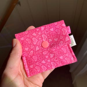 May include: A small, pink fabric pouch with a repeating heart pattern. The pouch is secured with a matching pink button. The fabric has a light pink background with darker pink hearts. A small tag is attached to the side.