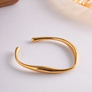 May include: A gold-colored, open cuff bracelet with a smooth, flowing design. The bracelet has a curved shape with rounded ends, and it sits on a white surface. The jewelry piece reflects light, highlighting its polished finish.