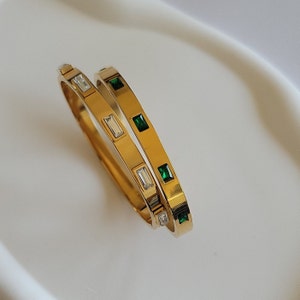 May include: Two gold bangle bracelets with green and clear rectangular stones. The bracelets are stacked on top of each other.