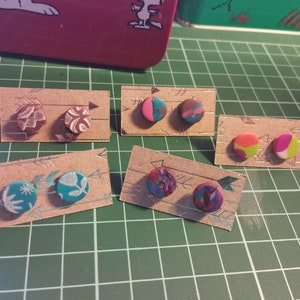 Könnte beinhalten: Sechs Paare kleiner, runder, bunter Ohrringe, die auf braunen Karten mit Pfeilen präsentiert werden. Die Ohrringe sind aus Polymer Clay gefertigt und haben verschiedene Muster und Farben, darunter Pink, Türkis, Grün und Braun.