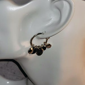 May include: A gold-colored hoop earring with multiple small, round, dark brown charms dangling from the bottom. The earring is attached to a white ear model, showcasing the design and style of the jewelry.