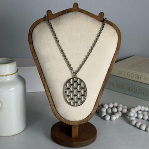 May include: A silver-toned necklace with a geometric oval pendant displayed on a wooden jewelry stand. The pendant features a repeating pattern of oval shapes. The necklace is on a cream-colored background.