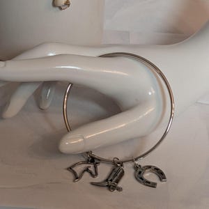 May include: A silver-colored bangle bracelet with three charms: a horse head, a cowboy boot, and a horseshoe. The bracelet is displayed on a white mannequin hand.