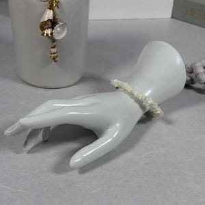 May include: A white ceramic hand figurine with a white bracelet. The bracelet is made of small, irregular white pieces. A white ceramic vase with a gold charm is in the background. The background is a light gray surface.