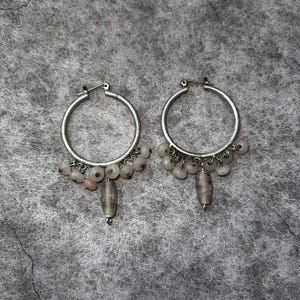 May include: A pair of silver hoop earrings with dangling beads. Each earring features a silver hoop with small, light pink and white beads hanging from the bottom. A larger, gray and white striped bead dangles from the center of each earring.