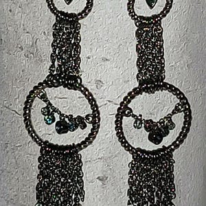 May include: A pair of dark gray dangle earrings. Each earring features a small green gem at the top, a twisted metal ring, and a cascade of chain with small, dark beads. Additional small gems are attached to the ring.
