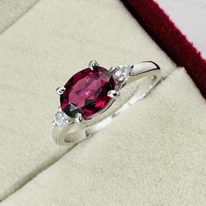 May include: A silver ring with a large oval-shaped red gemstone and two smaller round white gemstones. The ring is in a red velvet box.
