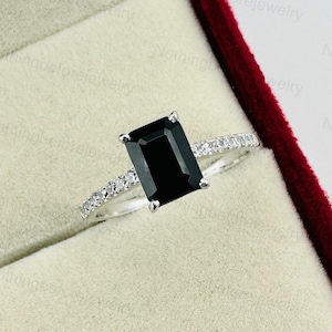 Puede incluir: Un anillo de plata con una piedra preciosa negra de talla esmeralda y una banda de pequeños diamantes.