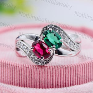 Peut inclure: Bague en argent ornée de deux pierres précieuses, une émeraude verte et un rubis rouge, serties dans un motif tourbillonnant. La bague est ornée de petites pierres claires et présentée sur un écrin de velours rose.