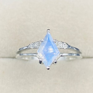May include: A silver ring featuring a kite-shaped, iridescent gemstone. The gemstone has a pale blue hue with a subtle shimmer. The ring band splits into two strands, with small, decorative accents on either side of the gemstone.