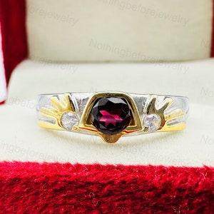 May include: A gold and silver ring with a large, dark red gemstone in the center. The ring has two smaller white gemstones on either side of the main stone.