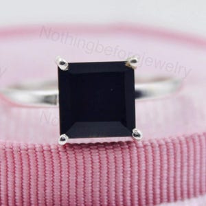 Könnte beinhalten: Ein silberner Ring mit einem großen, quadratischen, schwarzen Edelstein. Der Ring liegt auf einer rosa, bandartigen Oberfläche. Der Edelstein ist der Mittelpunkt mit einer polierten, reflektierenden Oberfläche.