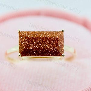 May include: A gold ring featuring a rectangular, sparkling, brown gemstone. The ring is set against a soft pink background, highlighting the jewelry's design. The band is a simple, polished gold.