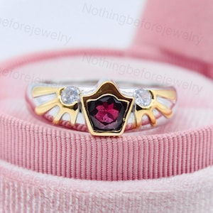 May include: A gold and silver ring with a red gemstone and two clear gemstones. The ring is in a pink velvet ring box.