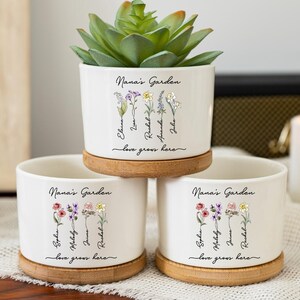 Könnte beinhalten: Drei weiße Keramik-Blumentöpfe mit Bambus-Untersetzern, jeweils mit floralen Illustrationen und dem Text "Nana's Garden" und "love grows here." Ein Topf enthält eine grüne Sukkulente. Die Töpfe stehen auf einer Holzoberfläche.