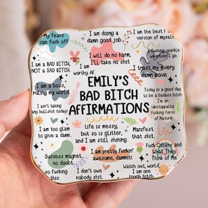 Könnte beinhalten: Ein weißer Keramikteller mit goldenem Rand, auf dem "Emily's Bad Bitch Affirmations" und verschiedene ermächtigende Sätze in Schwarz und Rosa stehen. Der Teller ist quadratisch mit abgerundeten Ecken.