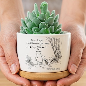 Peut inclure: Un petit pot de fleurs en céramique blanche avec une plante succulente verte. Le pot présente une illustration en noir et blanc de Winnie l'ourson et Porcinet, avec le texte "Never forget the difference you make Mrs. Gloria" et "Thank you Brenda". Le pot repose sur un dessous de verre rond en bois.