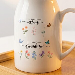 May include: A white ceramic pitcher with a handle, decorated with floral and butterfly decals. The text reads "First Mom, Now Grandma". The pitcher holds a bouquet of peach-colored flowers and greenery, resting on a wooden tray.
