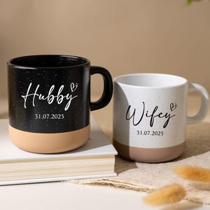 Könnte beinhalten: Zwei Keramikbecher, einer schwarz und einer weiß, mit beigefarbenen Böden. Der schwarze Becher trägt die Aufschrift "Hubby" mit einem Herz und dem Datum "31.07.2025". Der weiße Becher trägt "Wifey" mit einem Herz und demselben Datum.