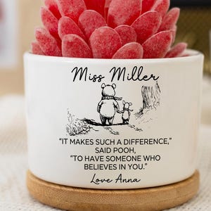 May include: A white ceramic pot with a red succulent plant. The pot has a Winnie the Pooh illustration and the text "Miss Miller". The quote reads "IT MAKES SUCH A DIFFERENCE, SAID POOH, TO HAVE SOMEONE WHO BELIEVES IN YOU." and "Love Anna".