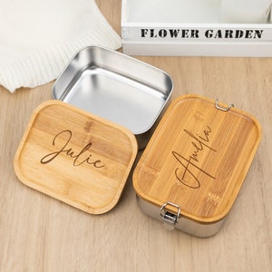 May include: Two stainless steel lunch boxes with bamboo lids. The lids are engraved with the names "Julie" and "Amelia".