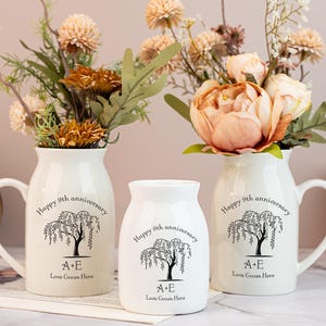 Puede incluir: Tres jarrones de cerámica blanca con asas y un jarrón pequeño, cada uno con un diseño de árbol negro y el texto "Happy 9th anniversary". Los jarrones contienen flores en tonos melocotón y marrón.