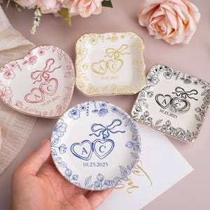 Op de afbeelding: Vier decoratieve ringschalen met bloemmotieven. Eén hartvormig, één vierkant en twee rond. Elke schaal heeft initialen, een datum en een strikdesign. De schalen zijn wit met gouden, roze, blauwe en zwarte accenten.