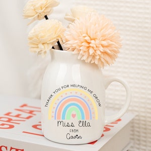 May include: White ceramic vase with a rainbow and the text "Thank you for helping me grow. Miss Ella from Gavin" on it. The vase is filled with dried flowers.