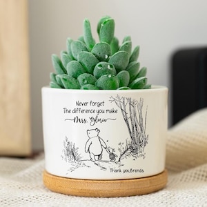 Könnte beinhalten: Ein weißer Keramik-Pflanztopf mit einer grünen Sukkulentenpflanze. Der Topf hat eine Winnie-the-Pooh-Illustration und den Text "Never forget the difference you make Mrs. Gloria. Thank you, Brenda." Er steht auf einem runden Bambussockel.