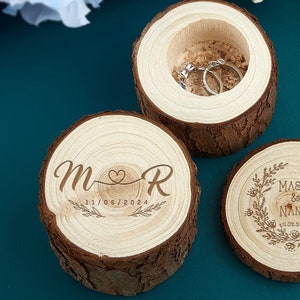 Op de afbeelding: Drie houten ringendoosjes. Eén doosje is open en onthult twee zilveren ringen. De andere twee doosjes zijn gesloten, met gegraveerde initialen en een datum op één, en namen en een datum op de andere. De doosjes zijn gemaakt van schijven van bomen.